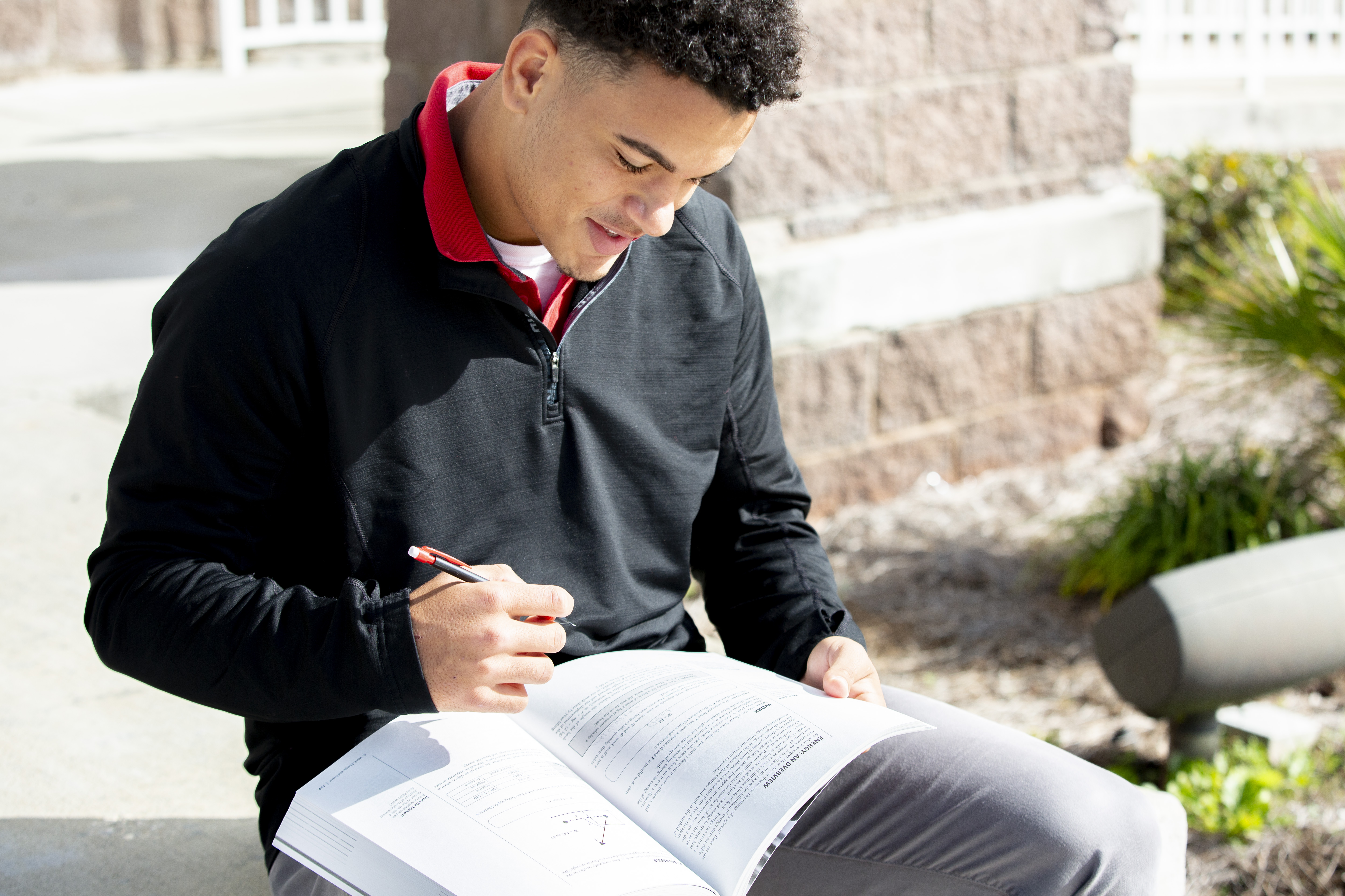 young male student is outside at school, looks down at his textbook with a pencil