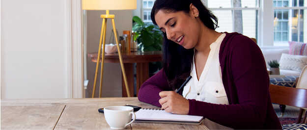 Young woman writing in a notebook at home with phone and coffee. How America Pays for Graduate School cover small version
